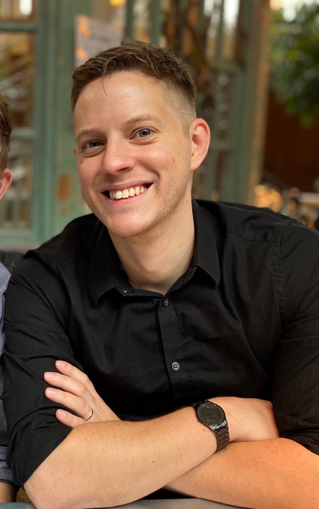 a young man (me) sat in a restaurant, arms crossed smiling and looking very happy. I wear a fitted black shirt, a think black metal watch and wedding ring. I look tanned. 